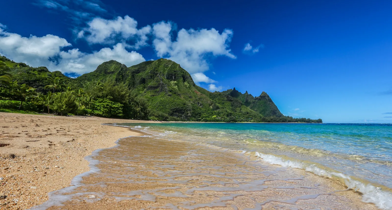 A bright North Shore beach with golden sand, clear turquoise water and a sharp green peak behind it.