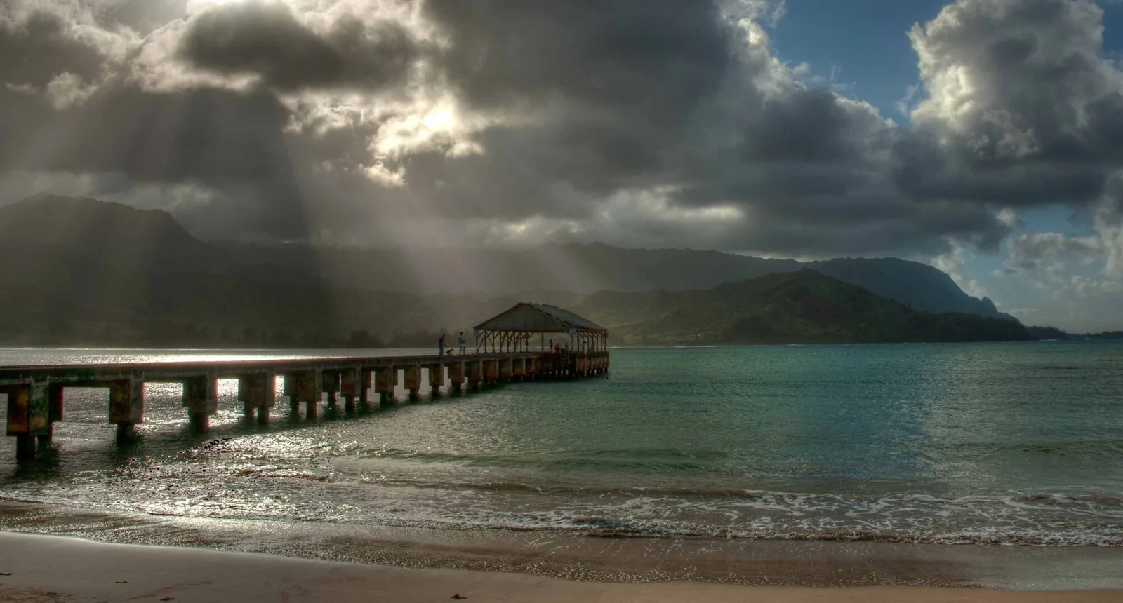Hanalei Pier reaching into Hanalei Bay at golden hour, sunbeams breaking through clouds over the green North Shore mountains.